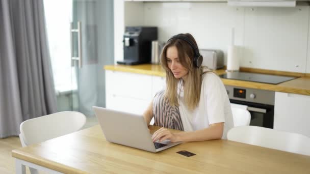 Woman in headphones at kitchen, listening to music and makes an online purchase using a credit card and laptop — Wideo stockowe