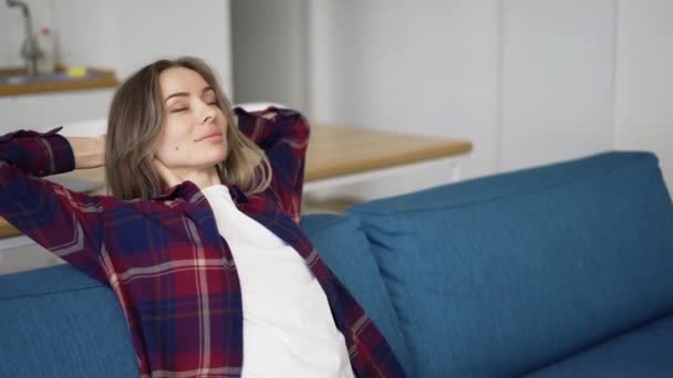 Ruhige junge Frau entspannt sich auf der Couch mit geschlossenen Augen hinter dem Kopf — Stockvideo