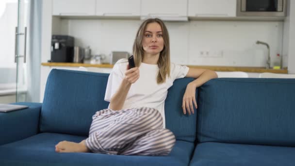 Una joven sentada en un sofá con un televisor de control remoto y canales de conmutación, vista frontal — Vídeos de Stock