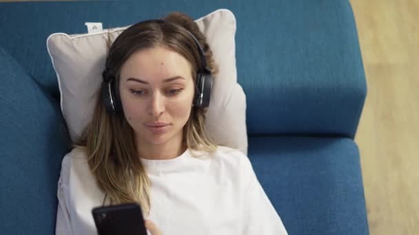 Kafkasyalı sakin sarışın kadın kanepeye uzanıp kulaklıkla müzik dinliyor. — Stok video