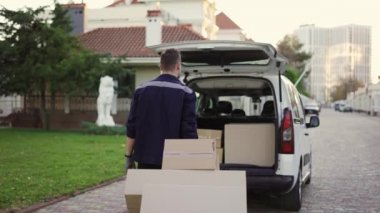 Young handsome smiling Caucasian delivery man with boxes on cart moving tovard to van with opened truck. Male post office worker Courier concept