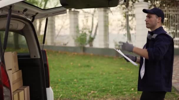 Handsome courier making notes in documents and takes cardboard box package out of delivery car. Courier in uniform, cap and gloves on the way to deliver postal parcel to a client — Vídeo de Stock