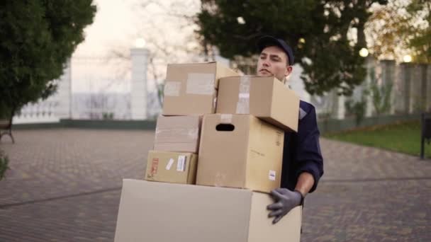 Hombre de entrega guapo en uniforme, gorra y guantes que llevan muchos paquetes de cajas de cartón pesadas al aire libre — Vídeos de Stock