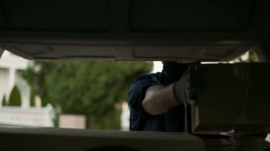Delivery service. Delivery man in a protective mask and gloves near a car putting cardboard box into the trunk. Man closing minivan trunk with parcels