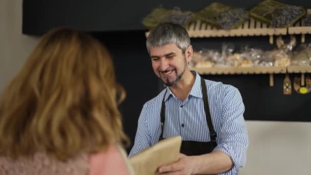 Friendly smiling male cashier in apron giving paper bag with bread to the woman. Female customer paying by phone and leave bakery shop happy — Video