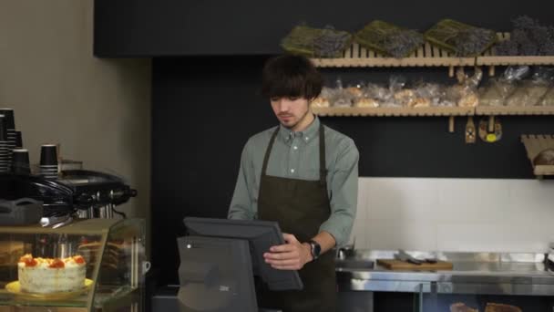 Cajero joven en delantal escribiendo en la pantalla táctil en la panadería. Primer plano, interior, cámara lenta — Vídeos de Stock