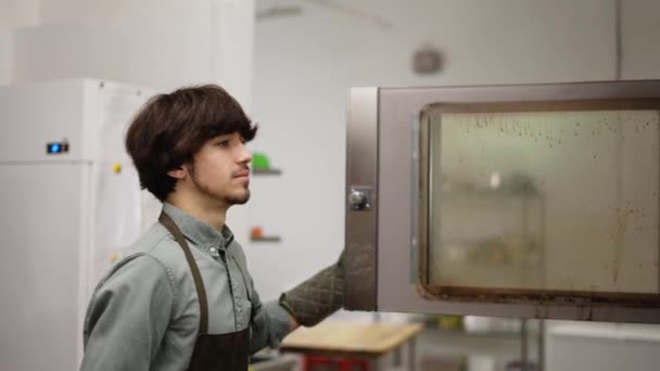 Young man baker taking fresh pastry out of oven in bakery kitchen. Side view of male chef opening oven and taking tray with tasty pies cooking in modern kitchen — Stock video