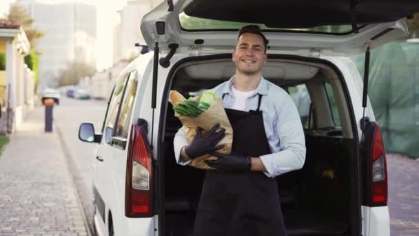 Happy delivery worker holding paper bag with food looking at the camera and smiling. Close up portrait of positive young man courier person in black apron and gloves — 비디오