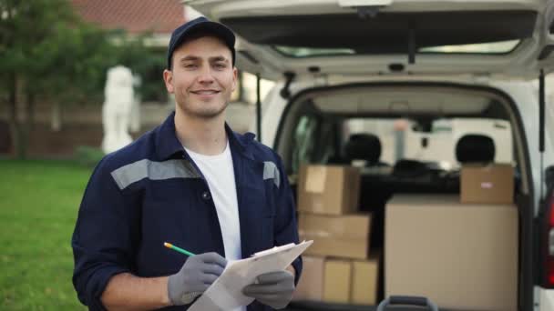 Happy smiling delivery service worker in uniform, cap and gloves makes notes on documents and standing on the street near the minivan. Close up footage — Video