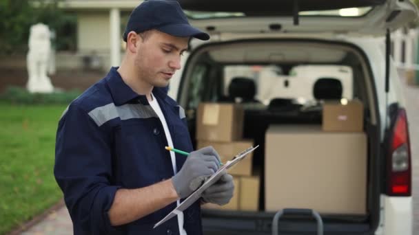 Young delivery man checking delivery list. Delivery man in cap and documentation in hands outdoor. Courier with parcel. Male handsome courier checks the parcels against the delivery list. Trunk with — стоковое видео