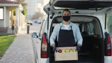 Friendly smiling man in mask holding wooden box with apples at street outdoors, portrait of happy deliveryman in apron and gloves standing near the trunk. Online shopping delivering, smart vegetables