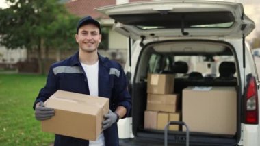 Happy smiling delivery service worker in uniform, cap and gloves holding paper box and standing near the minivan with opened trunk. Close up footage outdoor