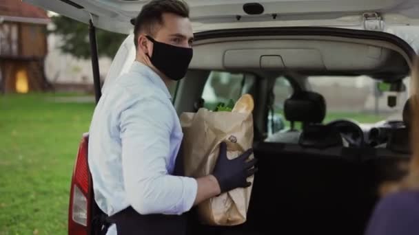 Female customer is getting paper bag of organic groceries from male courier in a mask, apron and gloves — Stockvideo