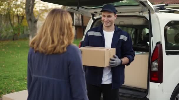 Woman meets a handsome friendly delivery man and takes a parcel box beside delivery van. Courier making notes about the parcel. Concept of courier, home delivery, online shopping — стоковое видео