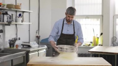 Handsome male baker sifting flour through a sieve at the kithen. Close up, indoor, slow motion