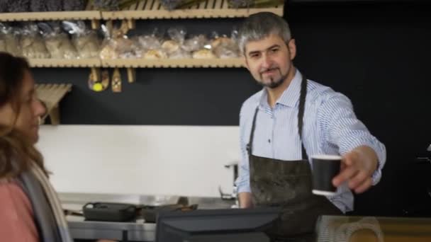 Όμορφος φιλικός αρσενικός barista με μαύρη ποδιά δίνοντας χάρτινο κύπελλο takeaway καφέ στη γυναίκα πελάτη. Εσωτερικός, φούρνος — Αρχείο Βίντεο