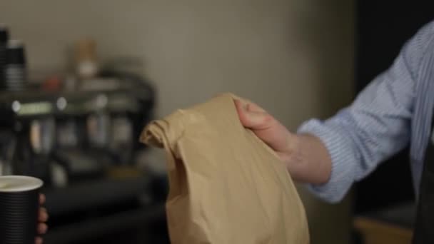 Male cashier in apron giving paper bag with bread to the smiling woman and she leaves holding paper cup with coffee. Close up, indoor, slow mo — Vídeo de Stock