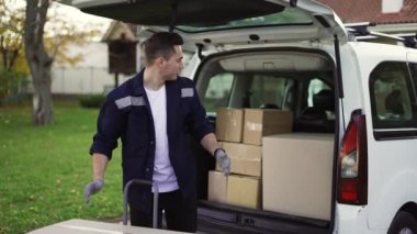 Handsome man takes boxes out of trunk and places them in a handcart. Parcel delivery, hand trolley