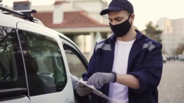 Handsome courier going out from the van and takes cardboard box package out of delivery car makes notes in delivery list. Courier in mask, cap and gloves on the way to deliver postal parcel to a