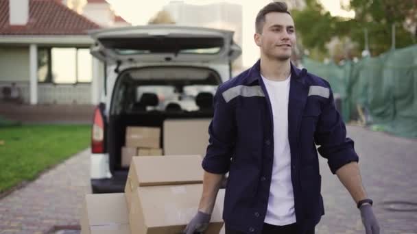 Young handsome smiling Caucasian delivery man with boxes on cart going from van with opened truck. Male post office worker Courier concept — стоковое видео