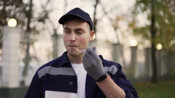 Relaxed courier in blue uniform , enjoying eating food from lunchbox and standing in autumn street near the cardboard boxes — Vídeo de Stock