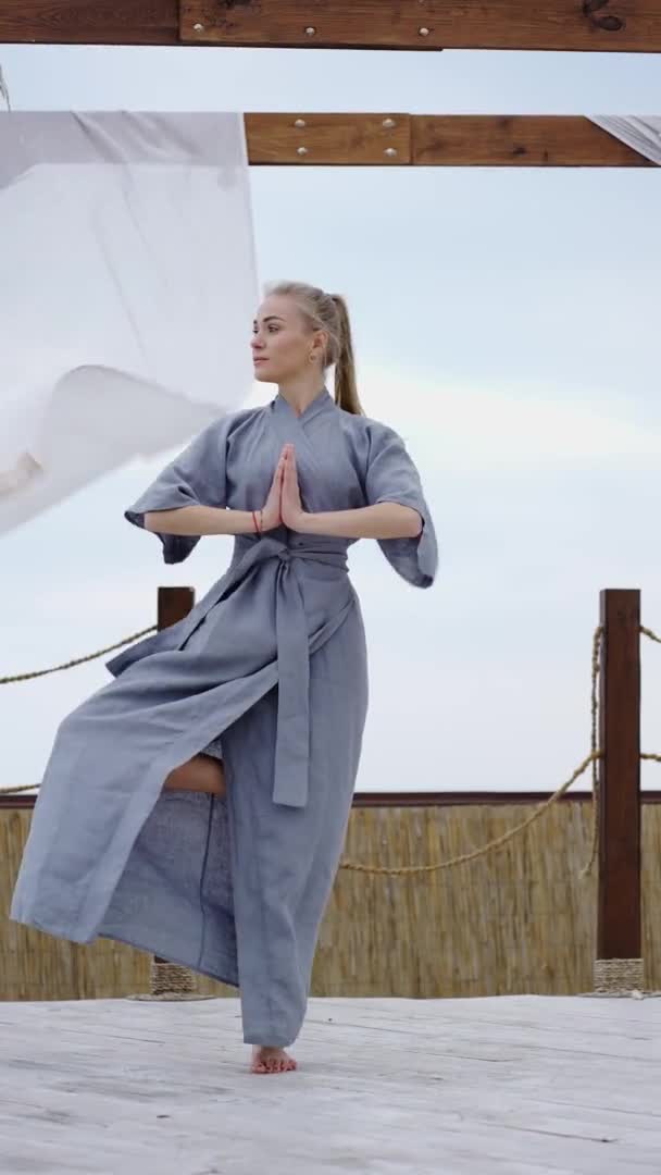 На терасі блондинка в блакитному халаті, що стоїть в позі йоги на ім'я — стокове відео