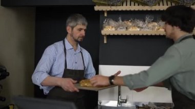 Handsome father in apron passes a tray of pastries to his young son and enters data into the bakery database. Indoor, close up, slow motion