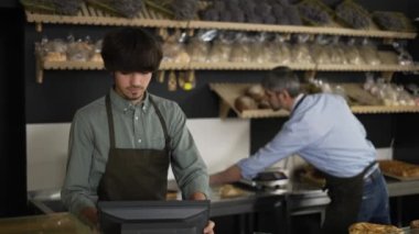 Young cashier in apron typing on touch screen, handsome bakery worker slicing bread in the background