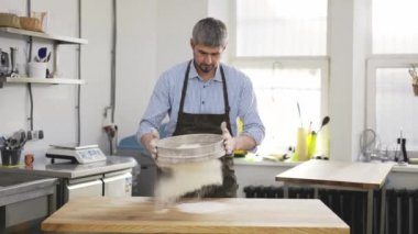 Handsome baker sifting flour through a sieve at the kithen. Close up, indoor, slow motion