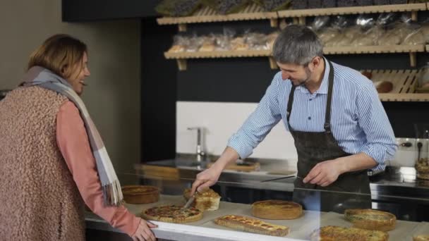 Vendedor de panadería feliz corta un pedazo de pastel para una cliente femenina y tienen una conversación casual — Vídeos de Stock