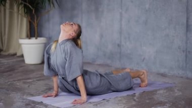 Şık cüppeli sarışın kadın minderde yoga yapıyor.