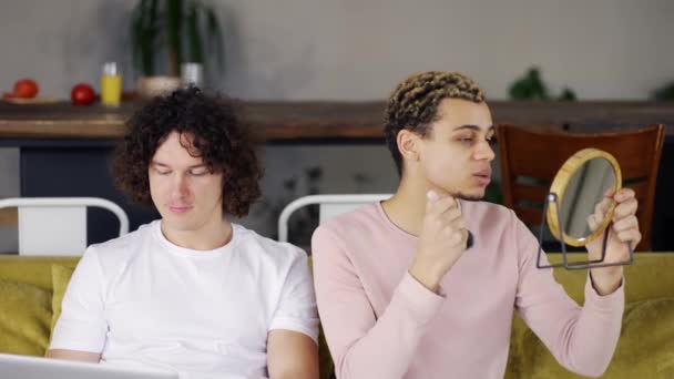 Два друга-гея сидят на диване, кудрявый парень делает макияж дома — стоковое видео