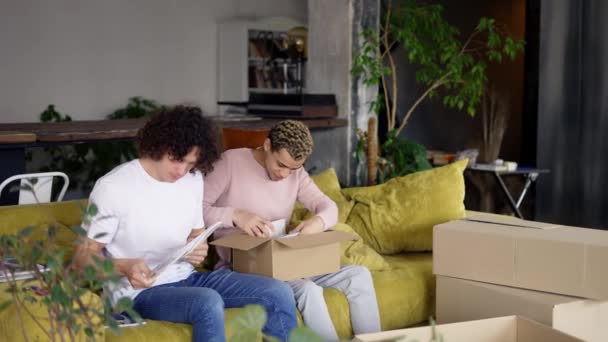 笑着的一对同性恋夫妇坐在沙发上搬到一个新公寓，打开盒子 — 图库视频影像