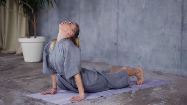 Blond vrouw in stijlvolle badjas doen yoga op mat — Stockvideo