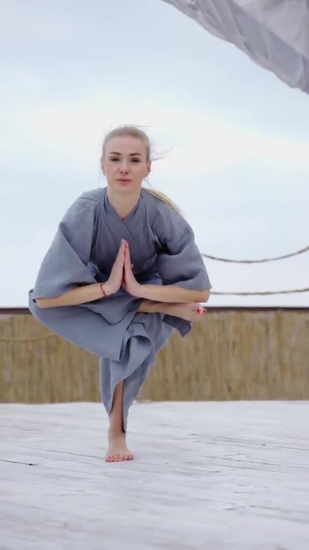 Привлекательная блондинка в халате занимается йогой Eka Pada Utkatasana на открытом воздухе — стоковое видео
