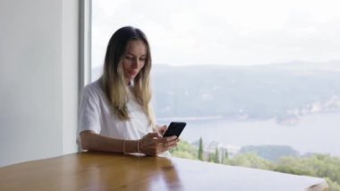 Kız, panoramik pencere manzarasıyla otururken telefonuyla internette sörf yapıyor.