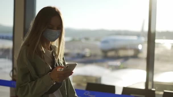 Blond vrouw in medisch masker maakt gebruik van telefoon lopen door luchthaven terminal — Stockvideo