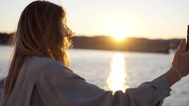 Blonde Frau blickt auf ihren Handybildschirm, während sie am Ufer steht und glücklich lächelt — Stockvideo