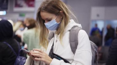 Havaalanı kalabalığında uçuş beklerken akıllı telefon kullanan maskeli kadın.