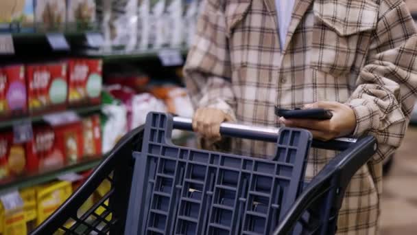 Onherkenbare vrouw met mobiel apparaat en trolley die producten koopt in het winkelcentrum — Stockvideo