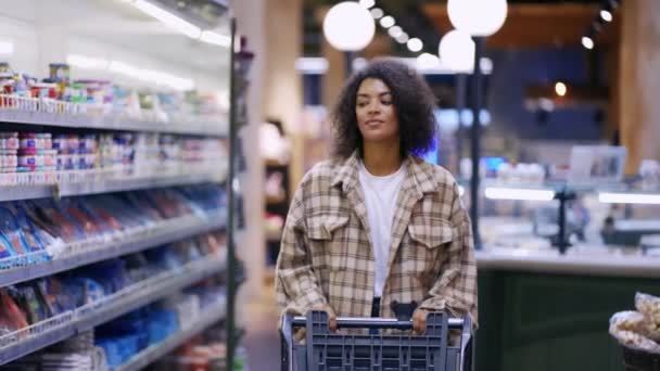 Uśmiechnięta Afroamerykanka przechodzi przez supermarket z wózkiem — Wideo stockowe