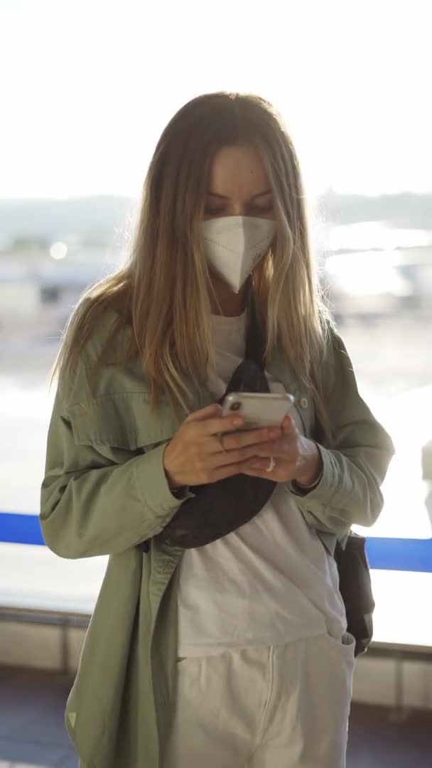 Νεαρή χαλαρή γυναίκα με μάσκα χρησιμοποιώντας το προσωπικό κινητό τηλέφωνο για συνομιλία με φίλους σε απευθείας σύνδεση, ενώ περιμένει την πτήση — Αρχείο Βίντεο