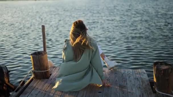 Ξανθιά κοπέλα κάθεται σε μια ξύλινη προβλήτα, πίσω όψη — Αρχείο Βίντεο