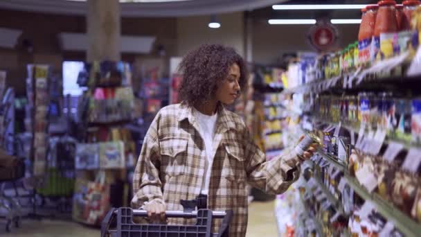 Piękna kobieta spaceruje po przejściu w supermarkecie z wózkiem sklepowym patrząc na półki — Wideo stockowe
