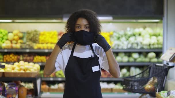 Retrato de una trabajadora afroamericana feliz parada en el supermercado, poniéndose una máscara de protección — Vídeo de stock