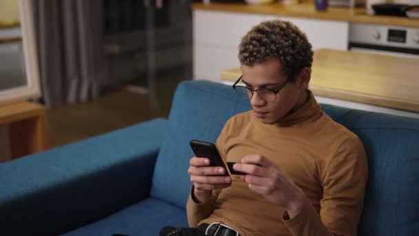Šťastný mladý muž nakupující on-line používá telefon a kreditní kartu na pohovce doma — Stock video