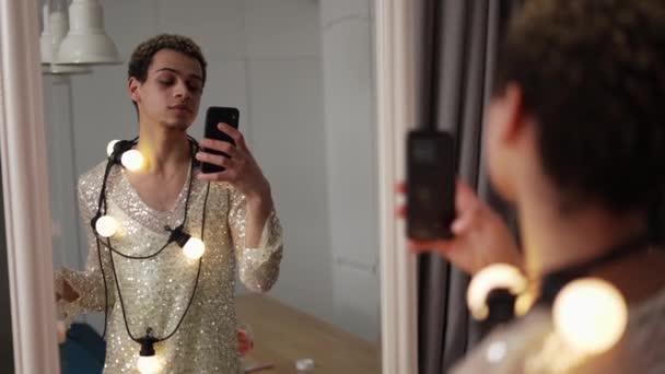 Un hombre en un vestido posando frente al espejo con guirnalda en el cuello y copa de vino usando el teléfono — Vídeos de Stock