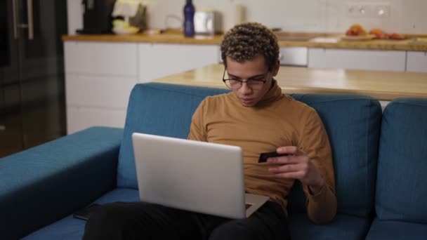 Joven compra en línea utiliza un ordenador portátil y una tarjeta de crédito en el sofá de casa — Vídeos de Stock