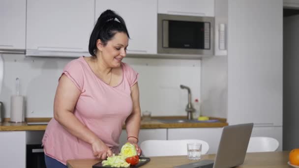 Jeune femme en surpoids cuisine salade, regarder la recette vidéo sur ordinateur portable en ligne — Video