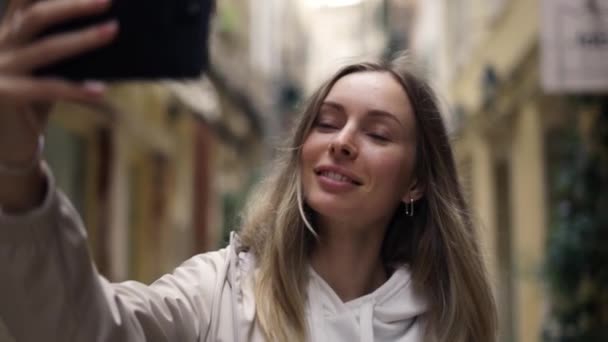 Блондинка в старом городе фотографирует на современный смартфон, делает автопортреты — стоковое видео